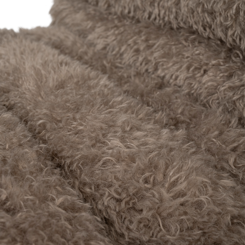 A lounge chair with visible rubber wood legs and a quilted and padded cushioned seat in a draped design. Made from a fluffy poodle fabric in the versatile colour taupe.