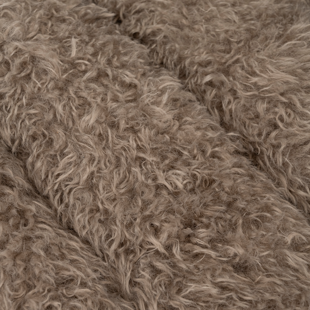 A lounge chair with visible rubber wood legs and a quilted and padded cushioned seat in a draped design. Made from a fluffy poodle fabric in the versatile colour taupe.